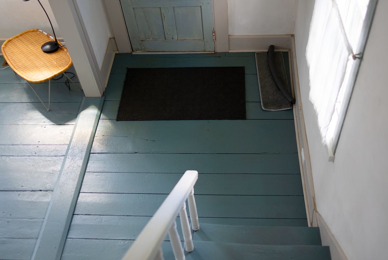 Blue-painted floorboards at the threshold — doormat, wicker chair, and lamp by the doorway