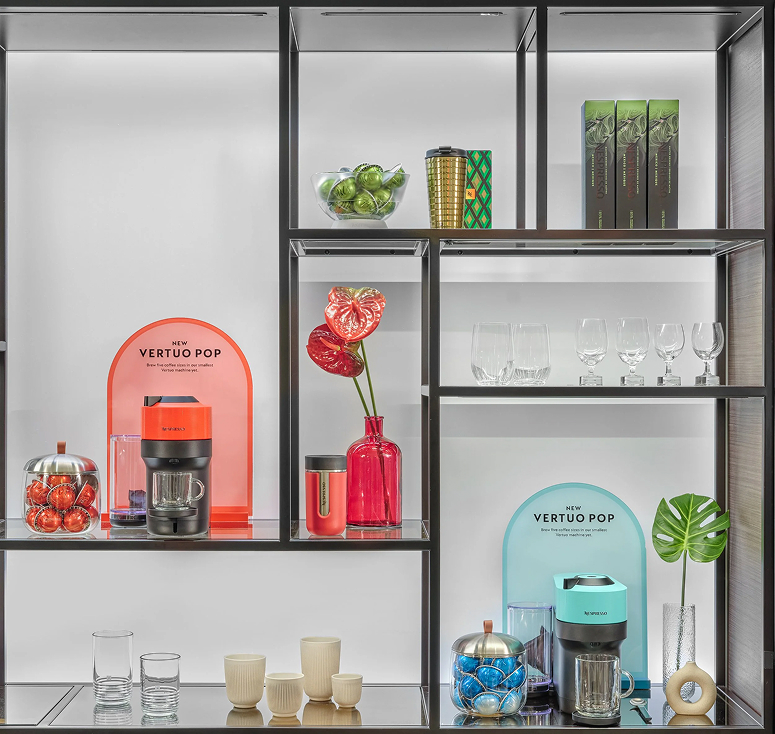 Boutique shelving — coral and teal arched campaign cards staging the Vertuo Pop machine alongside capsules and accessories